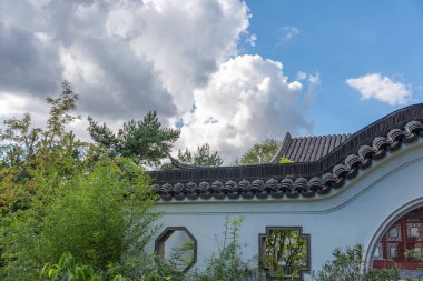 Yeşil bitkilerle çevrili, Çin tarzı çatılı geleneksel bir duvar. Beyaz bulutlar gökyüzünde sürükleniyor.