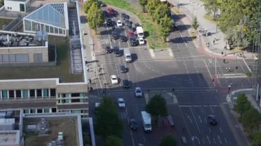 Trafik ışıklarında hareket eden arabalar, kamyonlar ve bisikletlerle dolu yoğun bir Berlin kavşağı. Yayalar, araçlar birden fazla şeritte sıralanırken geçitlerde bekliyorlar..