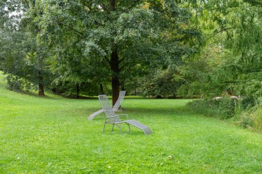 Çimenli bir çimenlikte iki modern metal şezlong duruyor. Uzun ağaçlar rahatlatıcı bir park atmosferi sağlar..