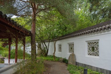 Çin bahçesinde kafes camlı beyaz bir duvar boyunca giden bir yol. Sakin sahneyi ağaçlar, bambu ve bir pavyon tamamlar..