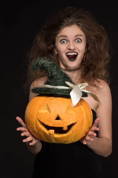 Beautiful girl with a gothic make-up as a witch smiles and holds an orange pumpkin on a black background for the holiday of Halloween