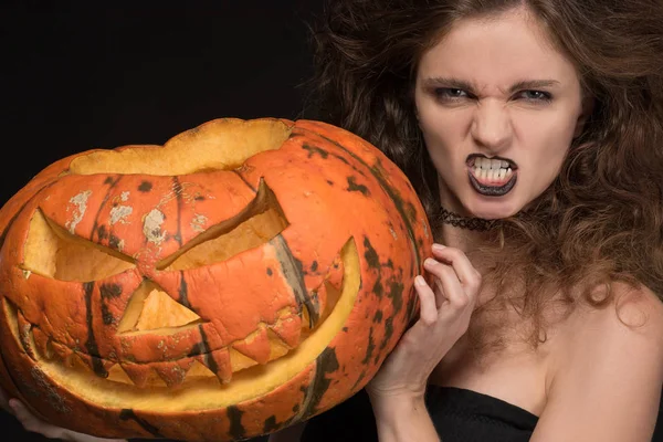 Beautiful girl with a gothic make-up as a witch smiles and holds an orange pumpkin on a black background for the holiday of Halloween