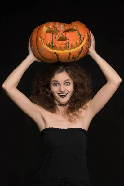 Beautiful girl with a gothic make-up as a witch smiles and holds an orange pumpkin on a black background for the holiday of Halloween