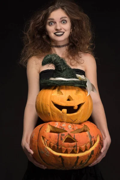 Beautiful girl with a gothic make-up as a witch smiles and holds an orange pumpkin on a black background for the holiday of Halloween