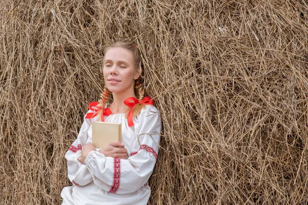 Beautiful blonde girl in a Slavic dress reads a book at a haystack in autumn
