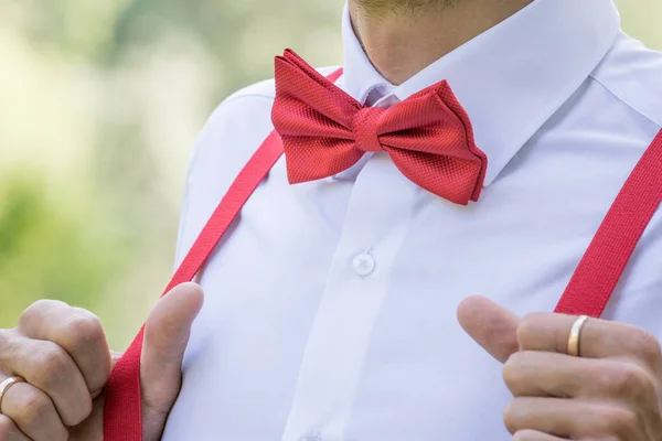 The groom in a white shirt with a red bow tie and in suspenders