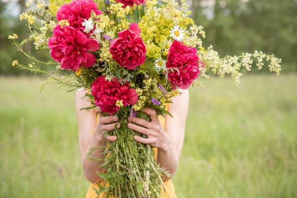 A girl in a yellow dress holds a large bouquet with peonies and wild flowers on a green meadow in summer