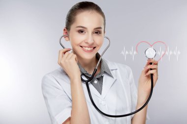 Genç kadın doktor kulaklara gri studio arka plan beyaz Tıbbi üniforma ve stetoskop giyiyor. Doktor işitme stetoskop ile