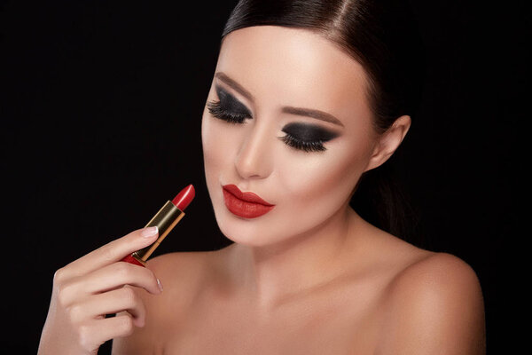 portrait of beautiful young woman with dramatic make up painting lips with red lipstick against black studio background, fake eyelashes