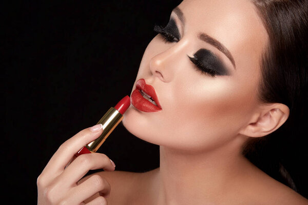 portrait of beautiful young woman with dramatic make up painting lips with red lipstick against black studio background, fake eyelashes