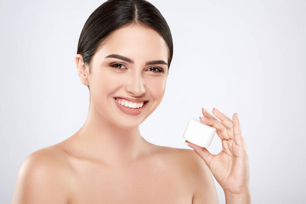 young smiling natural woman holding cream on skin against grey background