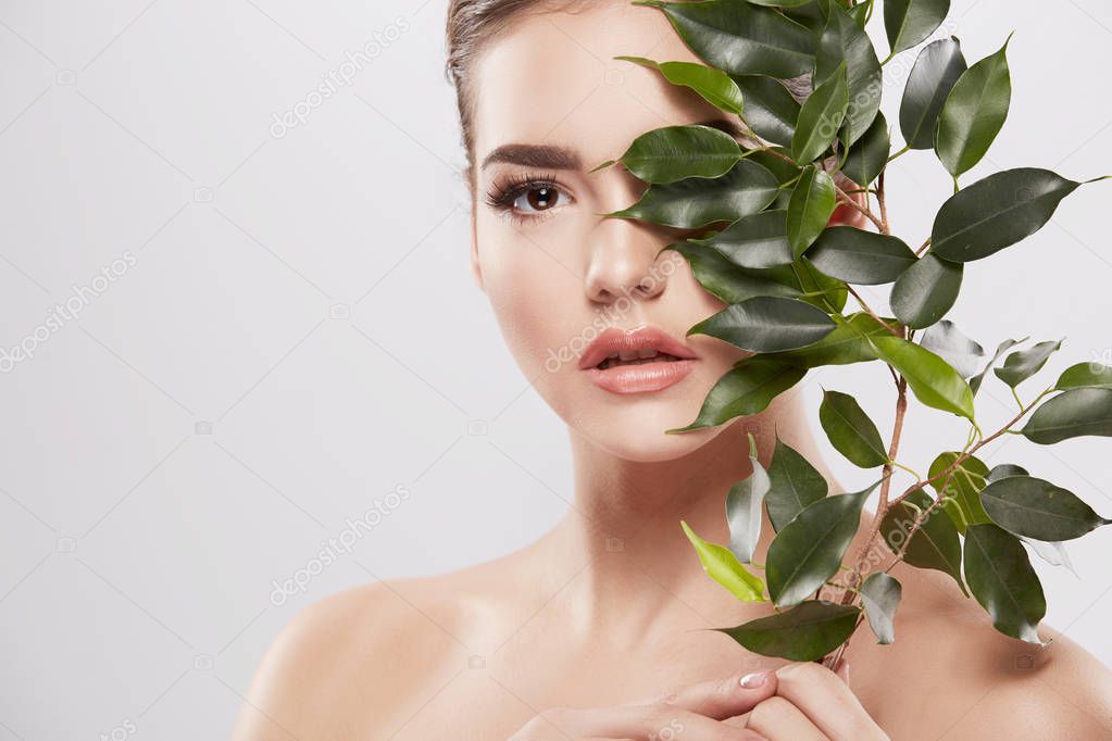 beautiful and natural young woman posing with green leaves at grey background, skin care concept, hydrated skin