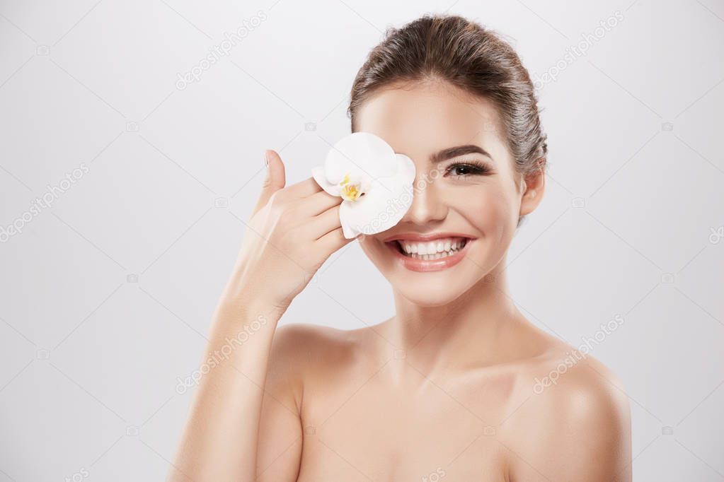 beautiful and natural young woman posing with white orchid flower at grey background, skin care concept, hydrated skin