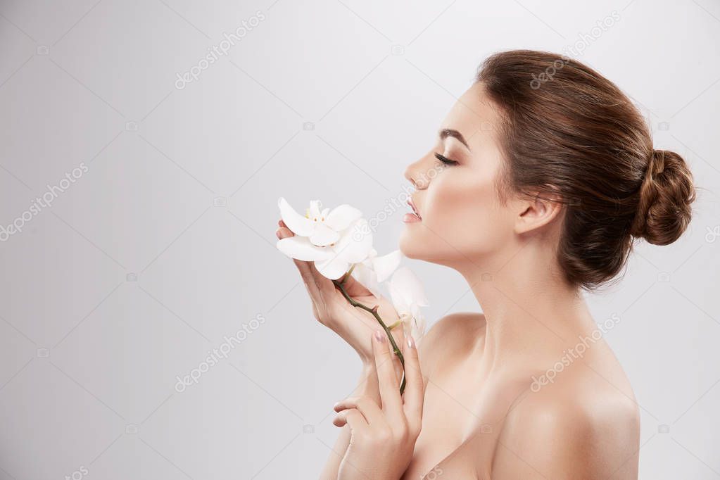 beautiful and natural young woman posing with white orchid flowers at grey background, skin care concept, hydrated skin