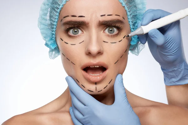 beauty portrait of scared woman, plastic surgery concept. Model in blue cap with puncture lines on face, hands in gloves drawing