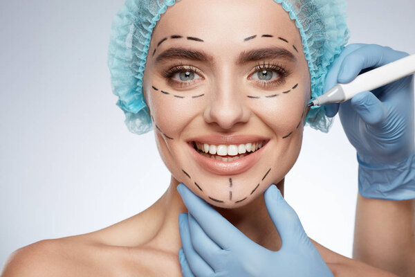 beauty portrait of smiling woman, plastic surgery concept. Model in blue cap with puncture lines on face, hands in blue gloves drawing