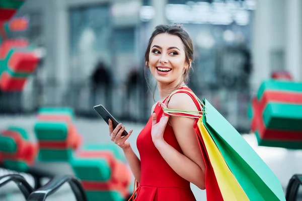 美丽的黑发年轻妇女与五颜六色的购物袋使用手机 网上购物的概念 — 图库照片