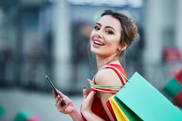 美丽的黑发年轻妇女与五颜六色的购物袋使用手机 网上购物的概念 — 图库照片