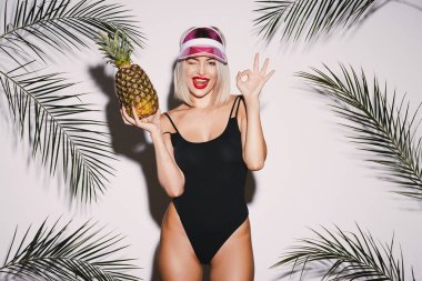 Ananas ile poz ile sarı saçlı giyen kız siyah takım elbise ve ile pembe kap ayakta beyaz stüdyo arka plan avuç içi, seksi modeli ile yüzme portre.