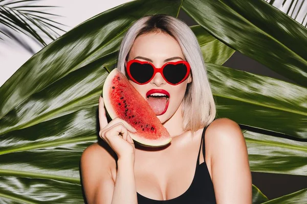 Arka plan yalama karpuz dilimi ve kırmızı dudaklar ile poz sıcak kız yüzme takım elbise ve güneş gözlüğü siyah sarı saçlı giyen ile ayakta üstünde palmiye yaprakları 