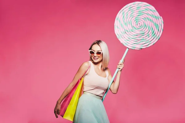 美丽的年轻女子戴着墨镜持有巨大的甜棒糖和五颜六色的购物袋站在粉红色的背景 面部表情 — 图库照片