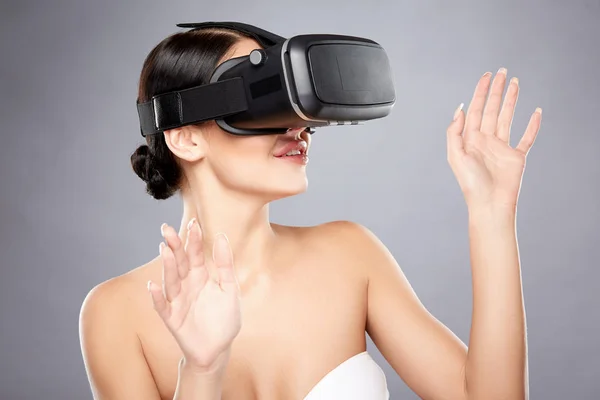Lovely girl with naked shoulders wearing virtual reality glasses at gray studio background, playing online game, virtual reality concept photo.