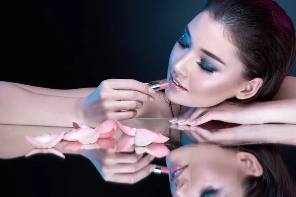 beautiful brunette woman with evening makeup, rose petals and lipstick on dark background