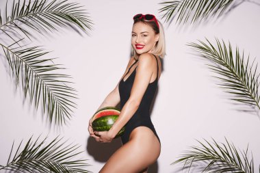 Kırmızı dudaklı, siyah mayo giyen, elinde karpuz tutarken arka planda palmiye yapraklarıyla poz veren genç bir kadın.