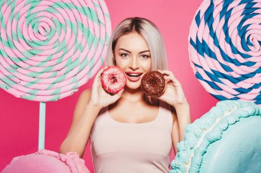 Pembe arka plan üzerinde çörek ve büyük tatlılar ile ayakta sarı saçlı çekici kız