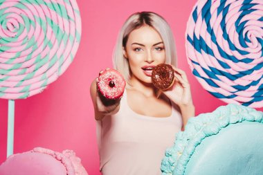 Pembe arka plan üzerinde çörek ve büyük tatlılar ile ayakta sarı saçlı çekici kız