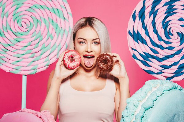 Attractive girl with blonde hair standing with doughnuts and huge sweets on pink background