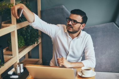 young handsome businessman in eyeglasses taking selfie on smartphone while working in cafe