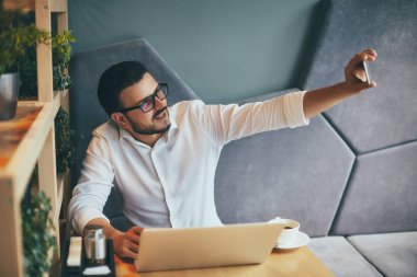 young handsome businessman in eyeglasses taking selfie on smartphone while working in cafe