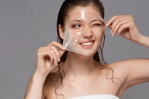 woman with wet hair peeling off face mask on grey background