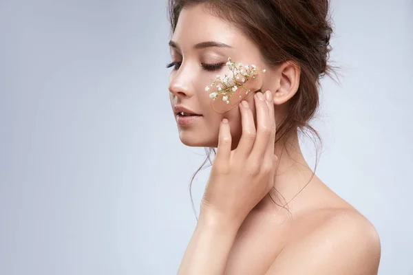 beautiful tender woman with flowers on face posing on grey backdrop