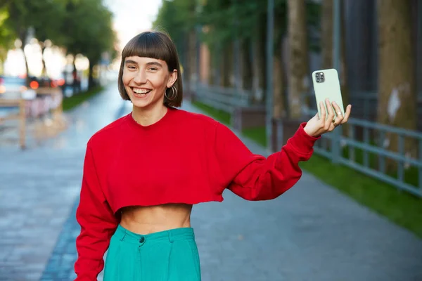 super happy millenial girl holding phone at the street, cute hipster woman in red stylish sweater having smartphone and looking to camera with smile