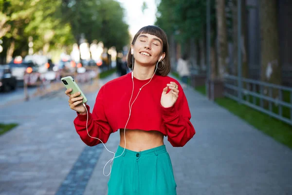 美しい女の子がイヤホンで路上に立って音楽を聴いたり かわいい千波万波の女性が赤いスタイリッシュなセーターを着てスマートフォンを持って音楽を聴いたり — ストック写真