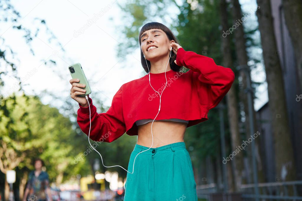 emotinal girl listening music on the street, cute millenial woman in red stylish sweater having using earphones and listen music