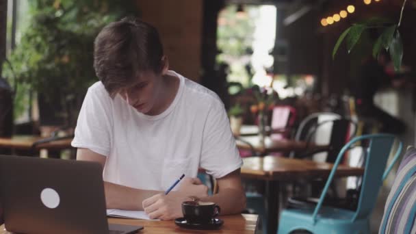 Ung student gör läxor i Café, frilansare med blyertspenna att rita på papper, modern arbetsplats sitter i kaféet. Öppen laptop i restaurang. — Stockvideo