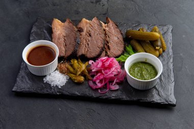 Dilimlenmiş et, siyah tabakta salatalık turşusu.