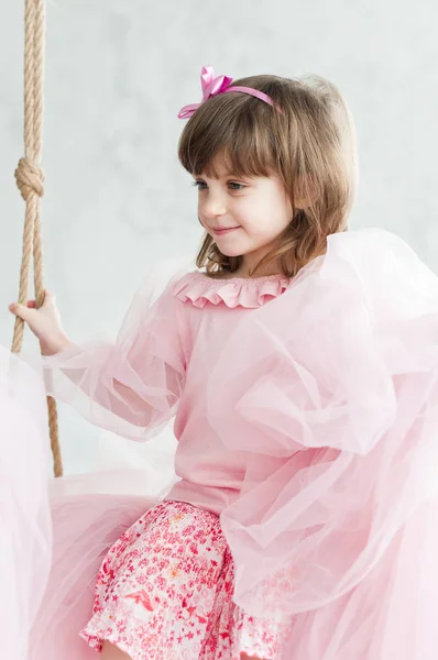 Cute little girl in pink clothes with bow on hair riding a swing indoors