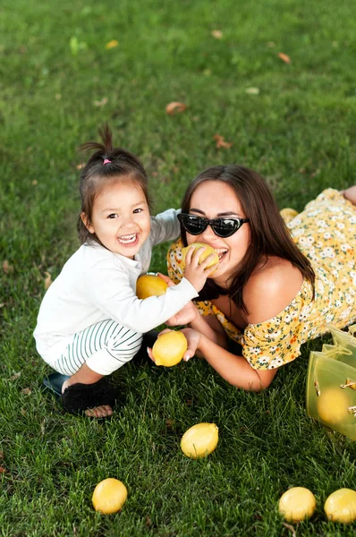 Funny mum with the daughter play with lemons lying on a grass in par