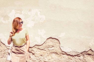 Yazın aşınmış bir duvarın yanında duran güneş gözlüklü kız.