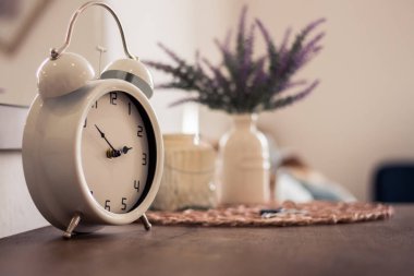 Beyaz, eski bir saatin perspektifli görüntüsü. Arka planda çan ve bitki var.