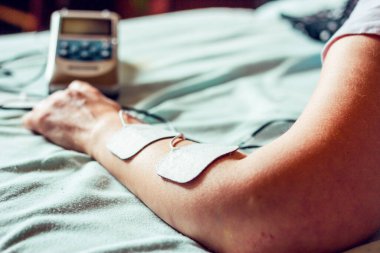 Bir kadının elinde elektrotlar eşliğinde ev kullanımı için küçük elektro uyarıcı. Masaj ve spor aletlerinde kullanılan elektrikli cihaz. Ağrı kesici ve kas geliştirme ekipmanları.