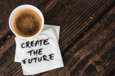 A full cup of coffee with a foam sitting on a white napkin with the message create the future handwritten on it placed on a wooden surface Concept image for inspirational messages clipart