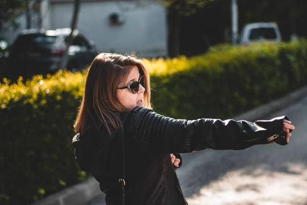 Esmer, deri ceket ve güneş gözlüğü takan genç bir kadın. Sokakta çılgınca davranıyor. Millennial gibi giyinmiş, dışarıda eğleniyor.