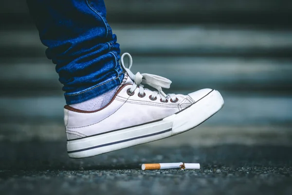 穿着白色运动鞋和牛仔裤的年轻人脚踩在破香烟上 坐在粗糙的柏油图上 整天不吸烟 戒掉有毒和危险的习惯 — 图库照片