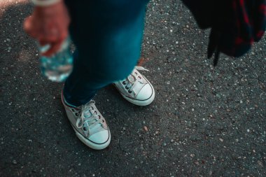 Beyaz spor ayakkabı ve kot pantolon giyen genç bir kadın elinde fotokopi alanı olan plastik bir şişe tutuyor. Gri asfalt üzerinde dikilen şık ayakkabıları olan sıradan giyimli bir milenyum.
