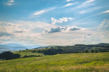 Ağaçlı yeşil tepelerin ve açık güneşli bir günde beyaz bulutlu mavi gökyüzünün güzel manzarası. Uçsuz bucaksız bereketli toprakların doğal ve kırsal manzarası.